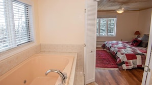 Un dormitorio con un ventanal grande, techo de madera, cama con colcha roja y blanca, y un baño con bañera.