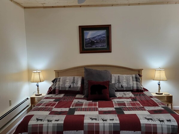 A bedroom with a wooden headboard, a quilt with animal prints, two bedside lamps, and a framed picture on the wall.