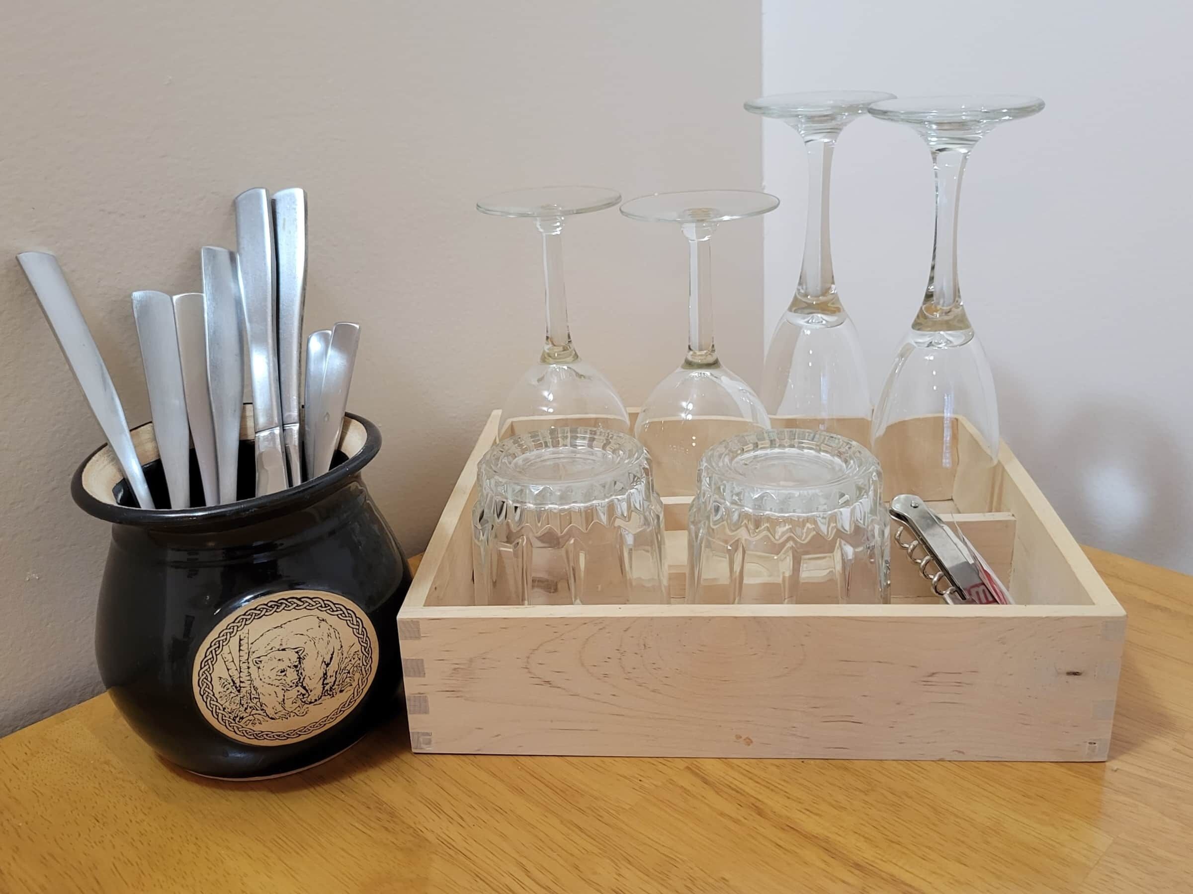 A wooden box containing wine glasses and a black mug with silverware.