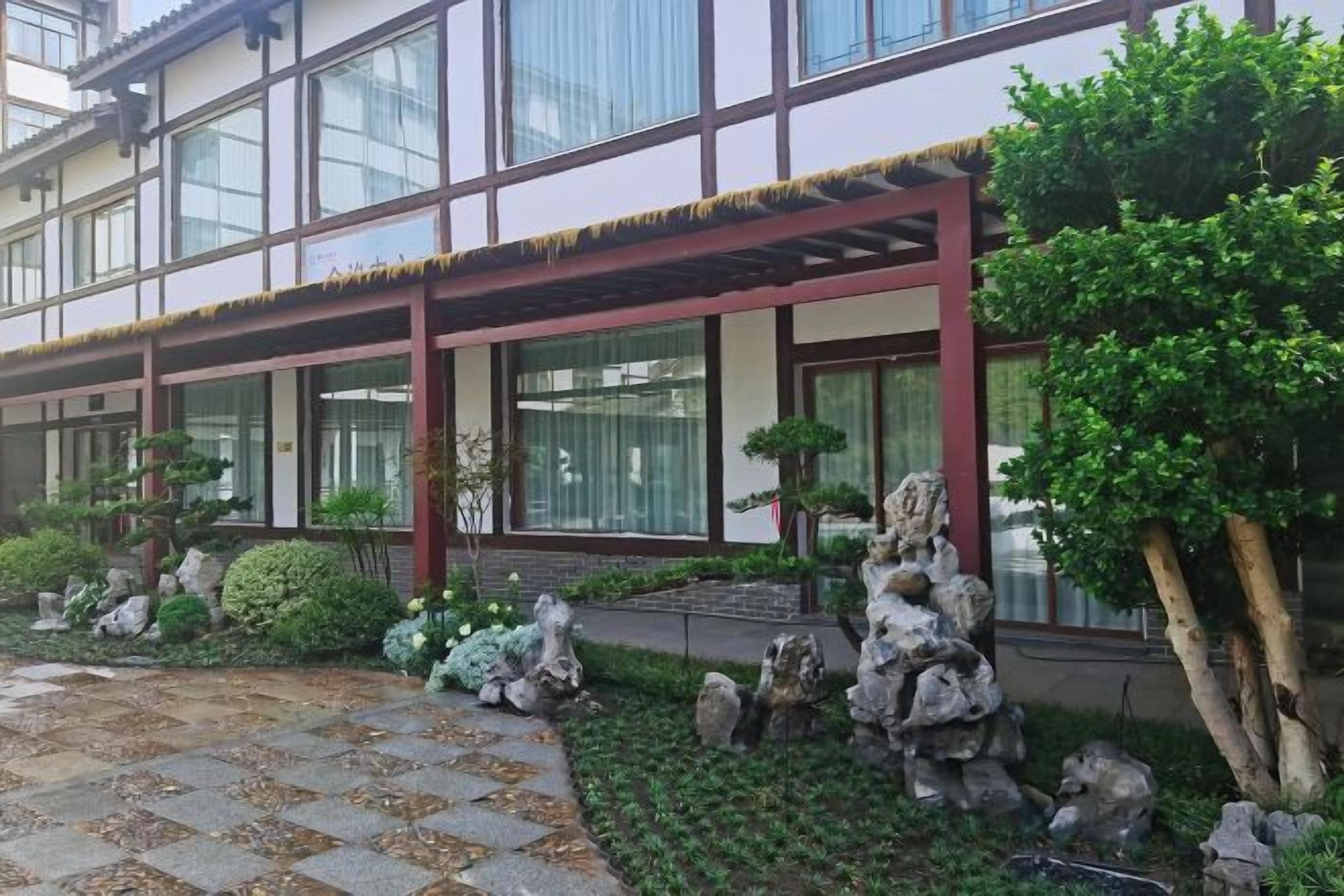 A building with a traditional architectural style, featuring a wooden structure with a tiled roof, large windows, and a well-maintained garden with rocks and plants.