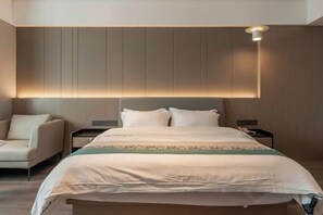 A modern hotel room with a large bed, a bedside table, a chair, and a wall-mounted light fixture.