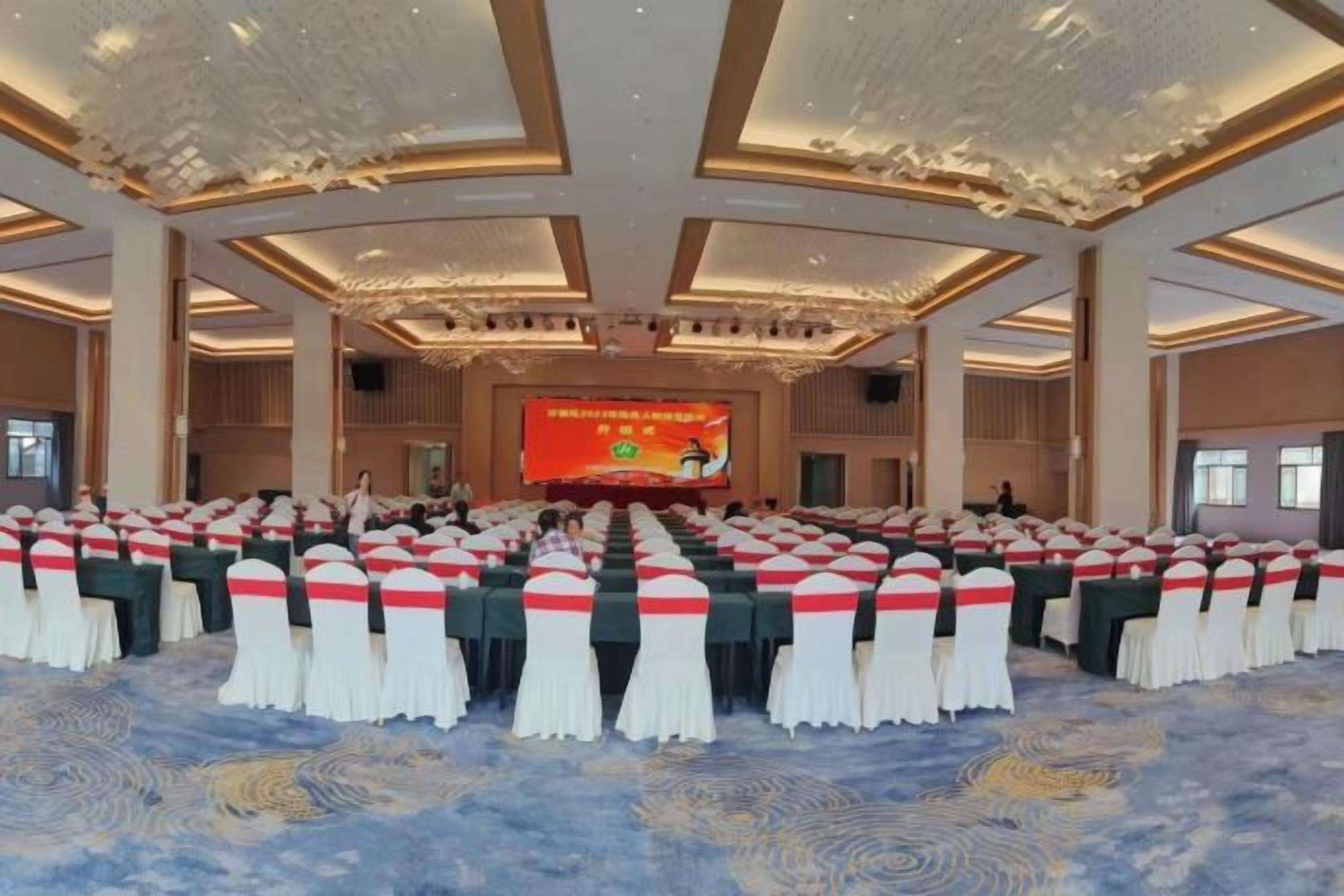 Un salón de conferencias con filas de mesas, un escenario con una pantalla y piso alfombrado.