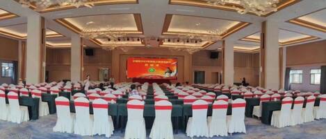 Un salón de conferencias con filas de mesas, un escenario con una pantalla y piso alfombrado.