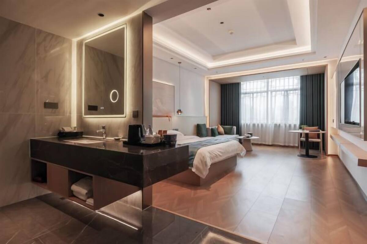 Una habitación de hotel moderna con una cama grande, un área de estar separada, un espejo amplio y un baño con lavamanos doble.