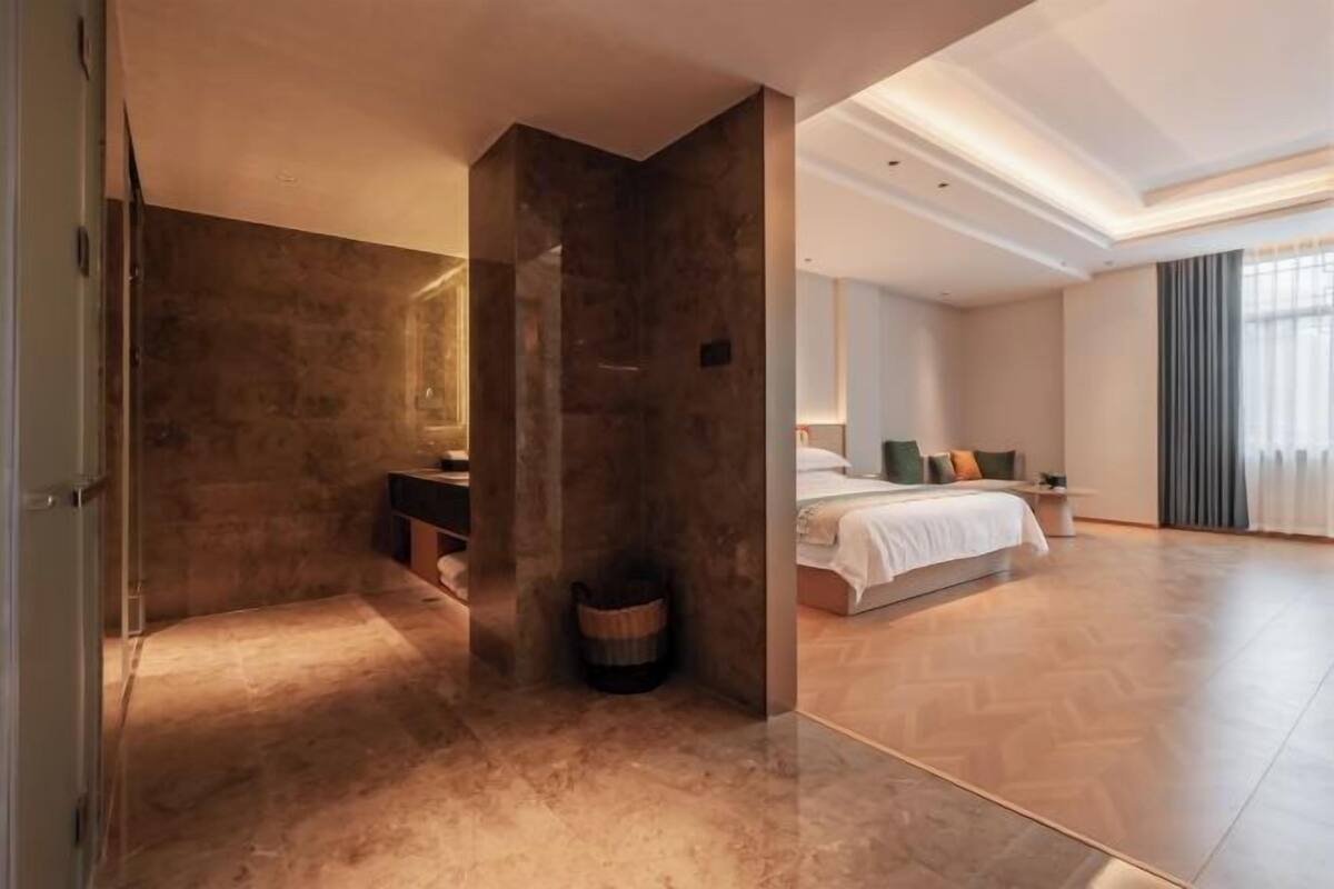 Una habitación de hotel moderna con una cama grande, una zona de estar con un sofá y una mesa de centro, y un baño con un espejo grande y un lavabo.