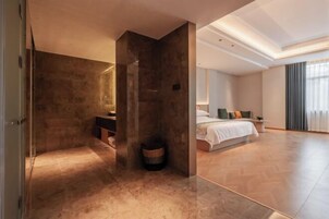 Una habitación de hotel moderna con una cama grande, una zona de estar con un sofá y una mesa de centro, y un baño con un espejo grande y un lavabo.