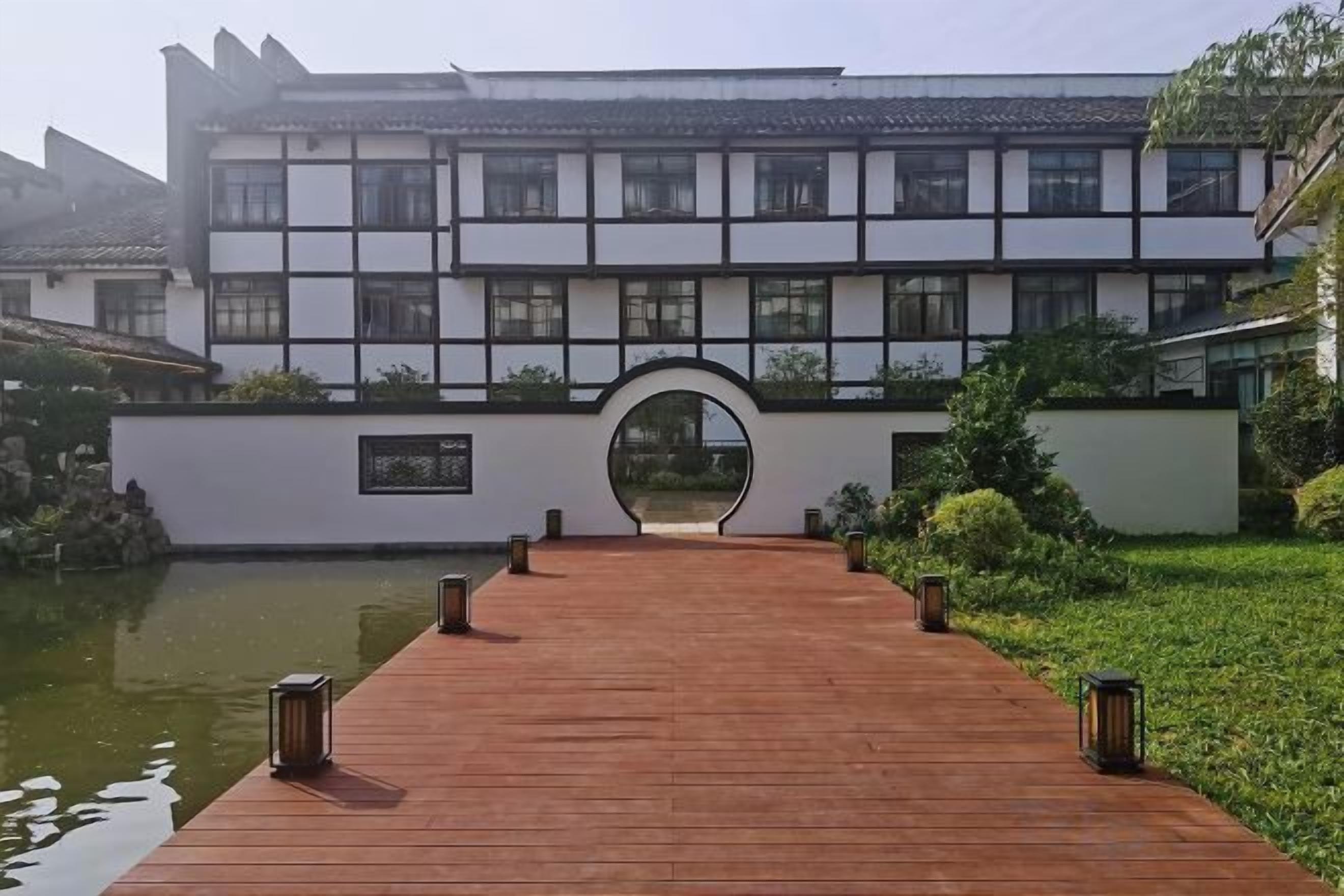 Edificio tradicional con pasarela de madera que conduce a una entrada arqueada, rodeado de vegetación y un cuerpo de agua.