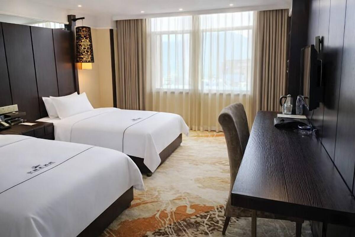 A hotel room with two beds, a desk, a TV, and a large window with curtains.