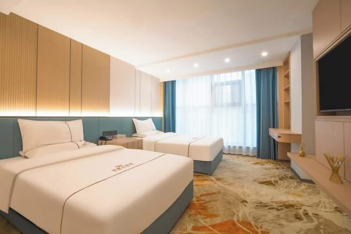 A hotel room with two beds, a TV, a desk, and a large window with curtains.