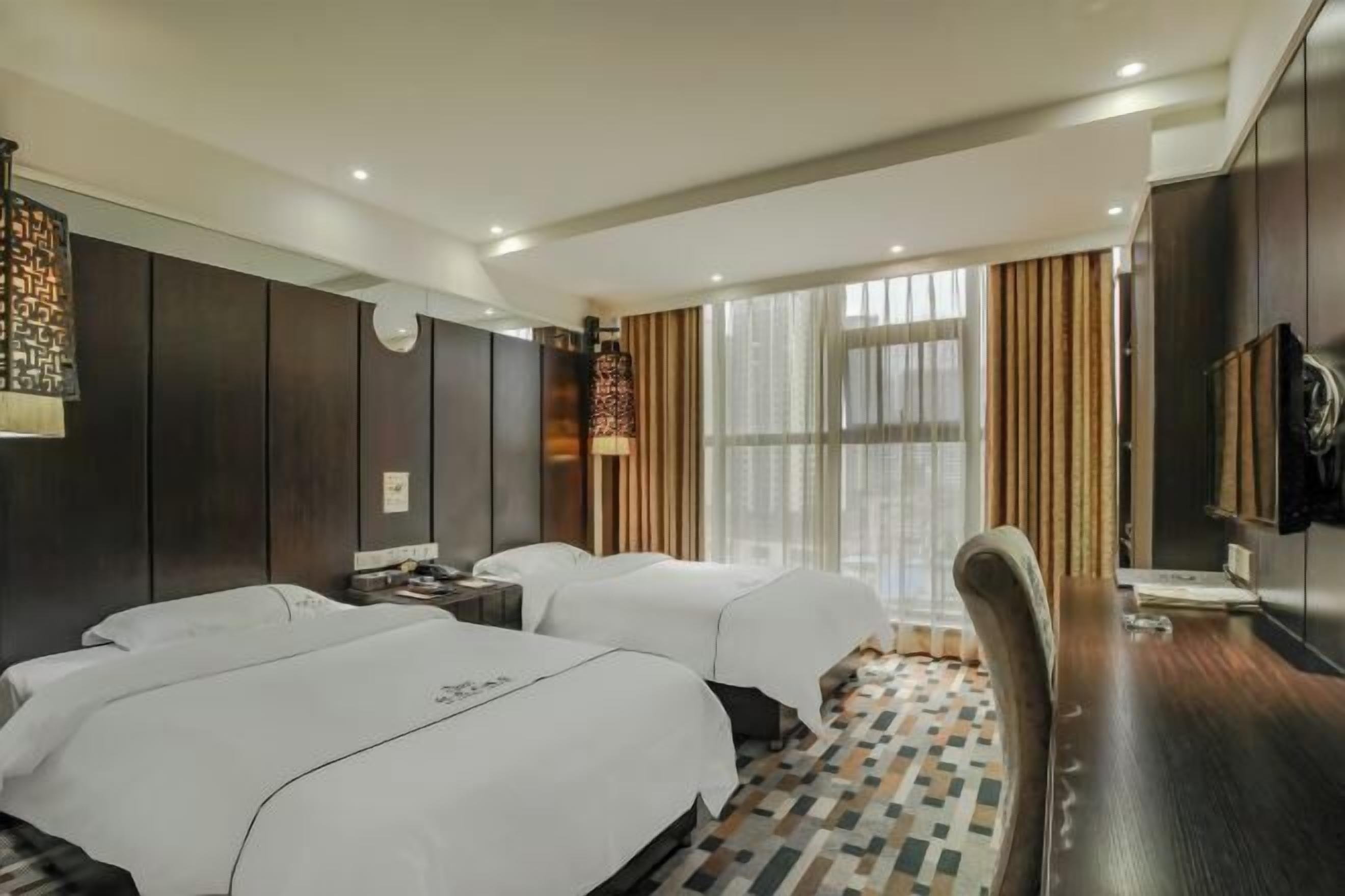 A hotel room with two beds, a desk, a chair, a TV, and a large window with curtains.