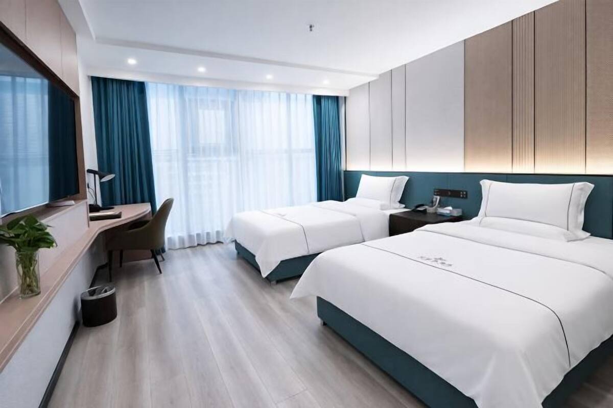 A hotel room with two beds, a desk, a chair, a lamp, and a plant.