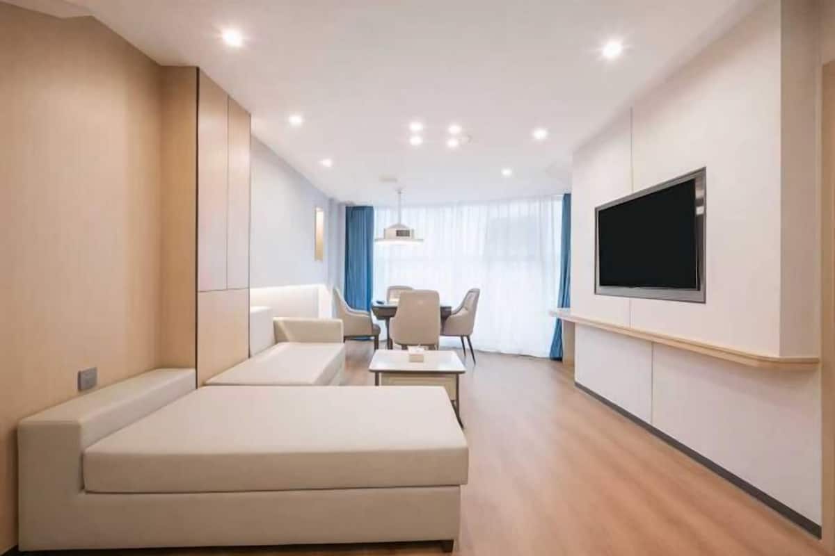 A modern hotel room with a sofa, a dining table with chairs, a flat-screen TV mounted on the wall, and a large window with curtains.