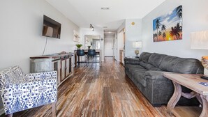 Una sala de estar moderna con un sofá gris, una mesa de centro de madera y un sillón estampado. Se ve un área de cocina con una mesa y sillas en el fondo.