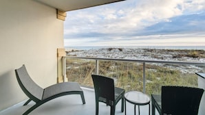 A balcony with two chairs and a small table, overlooking a beach with sparse vegetation and a cloudy sky. - Steps from Sugar-White Sand – Daily Beach Chairs Included (Pensacola Beach)
