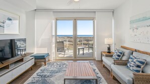 A modern living room with a large TV, a sofa, a coffee table, and a view of the beach through a sliding glass door. - Beach Chairs! Onsite Spa w/ Beach Club 1B Gulf Front (Pensacola Beach)