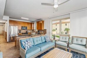 Une cuisine moderne avec des armoires en bois, un réfrigérateur en acier inoxydable et un four à micro-ondes encastré. Le salon est doté d’un canapé bleu avec des coussins à motifs et de deux fauteuils en osier.