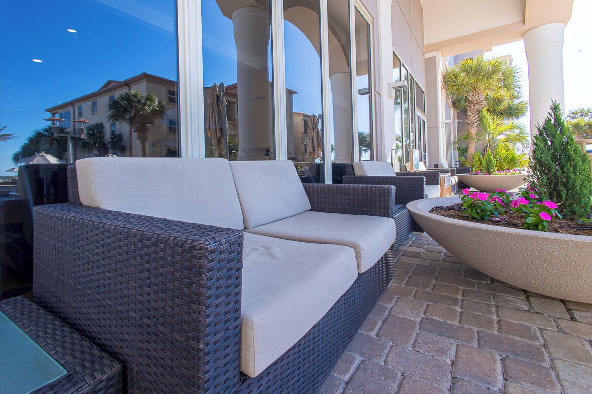 A wicker sofa with cushions, a potted plant with pink flowers, and a brick patio.