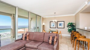 A modern living room with a sectional sofa, a dining area with a table and chairs, and a large glass door leading to a balcony with a view of the ocean.