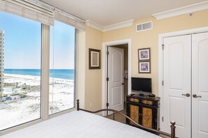 Ein Schlafzimmer mit großem Fenster mit Blick auf einen Strand, einem Bett, einer Kommode und einem Fernseher.