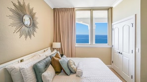 Une chambre d’hôtel avec un lit, un miroir, une fenêtre donnant sur l’océan et une porte.