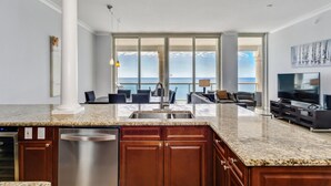 A modern kitchen with a granite countertop, stainless steel sink, and wooden cabinets. There is a dining area with a table and chairs, and a large TV on the right. The room has large windows offering a view of the ocean. - BC-2003 3B Beach Club Gulf Front /Daily Beach Chairs! (Pensacola Beach)
