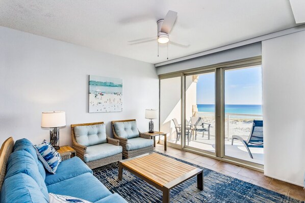 A spacious living room with a large window offering a view of the beach, a ceiling fan, and a comfortable seating area with a coffee table. - BC 204B 1B Gulf Front - Daily Beach Chair Set! (Pensacola Beach)
