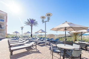 A sunlit outdoor seating area with lounge chairs and umbrellas, a brick patio, and a view of the beach and ocean.