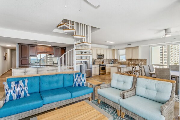 A spacious living area with a blue sofa, a light blue sofa, and a wicker chair. A wooden staircase leads to a kitchen area with modern appliances.