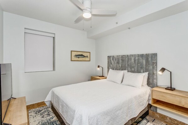 A bedroom with a bed, a ceiling fan, a framed picture on the wall, and a nightstand with a lamp.