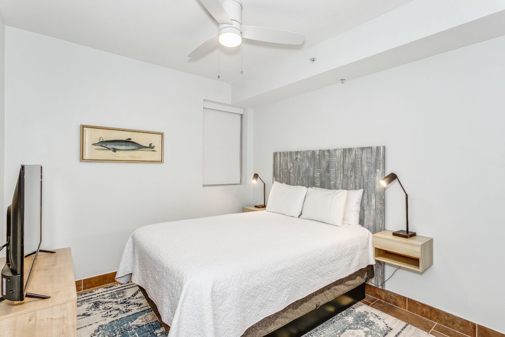 A bedroom with a bed, a ceiling fan, a framed fish picture, a nightstand, and a TV.