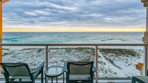 Ein Balkon mit zwei Stühlen und einem kleinen Tisch, mit Blick auf einen Strand mit Sand und spärlicher Vegetation.