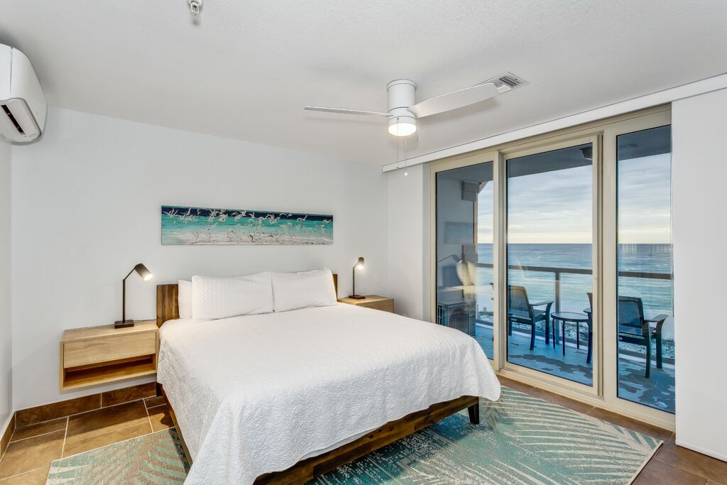 A bedroom with a bed, a ceiling fan, a painting, and a view of the ocean through large sliding doors.