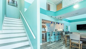 A modern interior with a staircase, a kitchen area, a dining space with bar stools, and a seating area with a flat-screen TV.