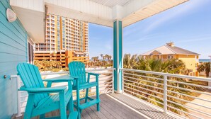 A covered outdoor area with turquoise Adirondack chairs and a table, overlooking a beach and a hotel building.