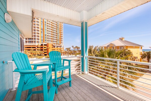 A covered outdoor area with turquoise Adirondack chairs and a table, overlooking a beach and a hotel building.