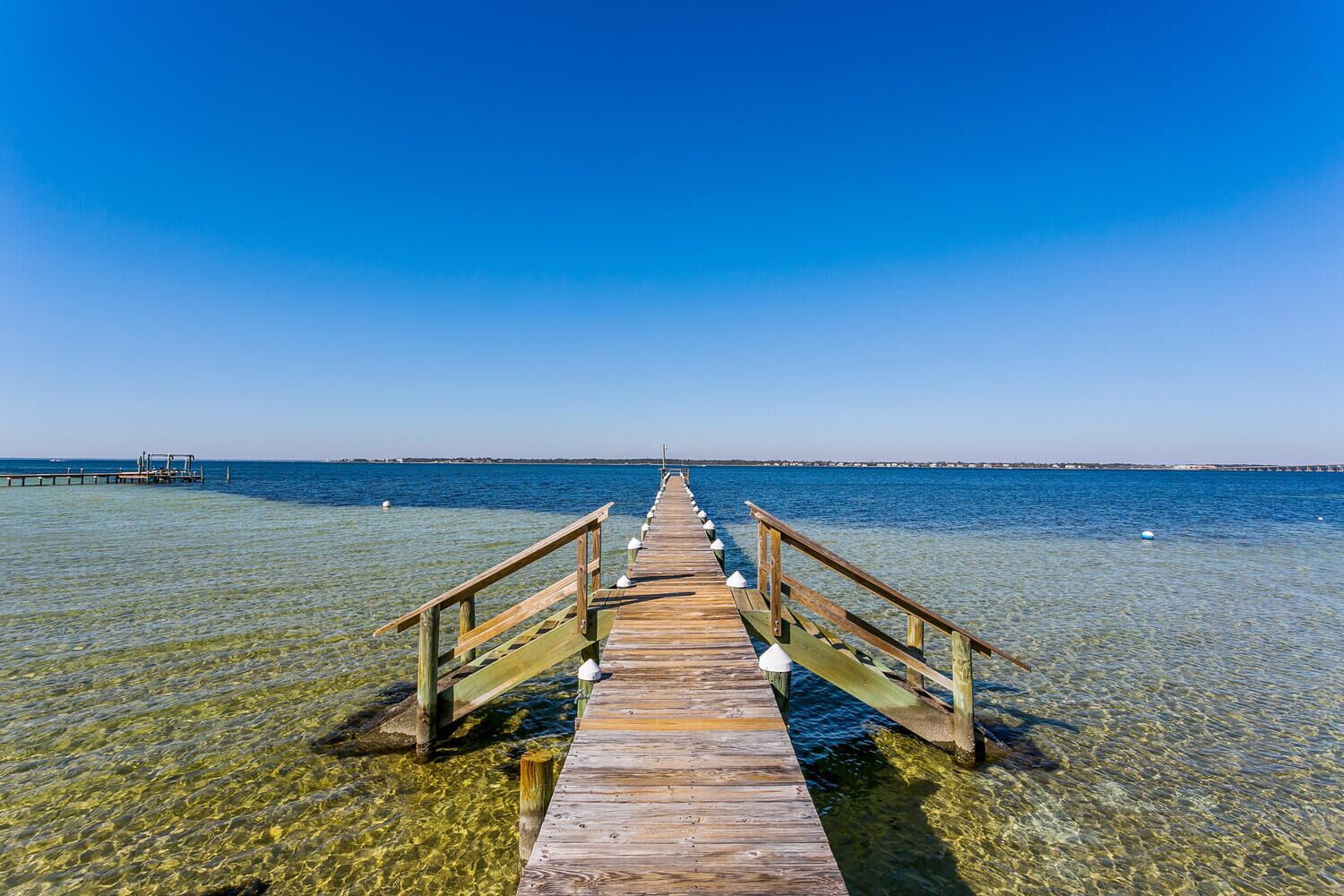 Eine hölzerne Anlegestelle erstreckt sich in ein klares, blaues Meer, im Hintergrund eine Küstenlinie.