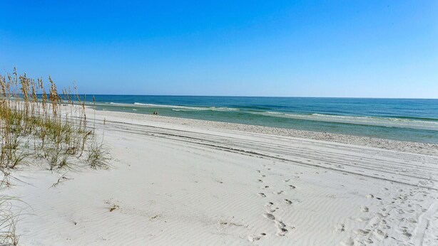 A beach with white sand, sparse vegetation, and footprints leading to the water. - Beach and Bay Views at Santa Rosa Dunes! (Pensacola Beach)