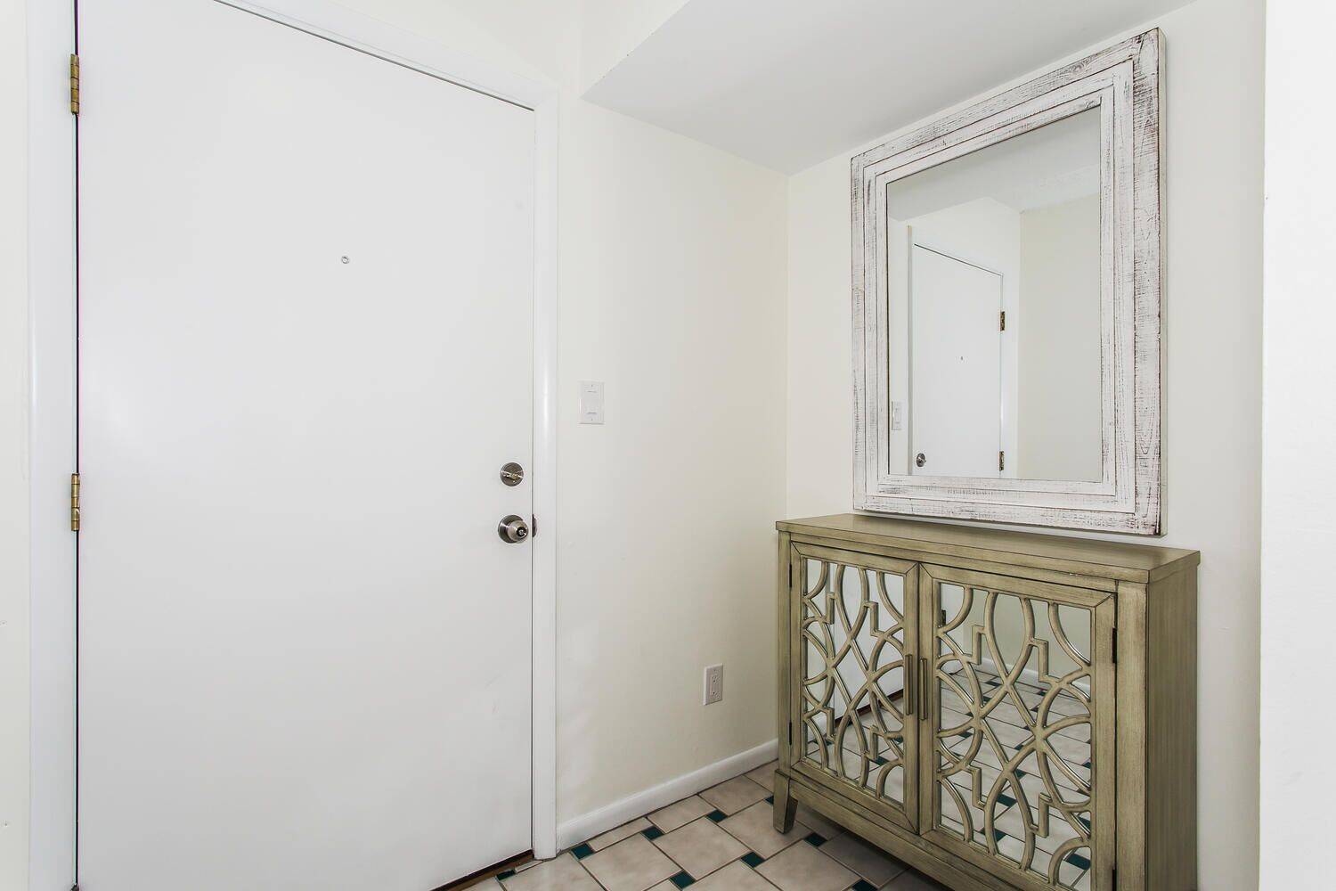 Uma porta branca com uma fechadura e um olho de chave, um espelho sobre um armário com um design geométrico, e um chão de azulejos.