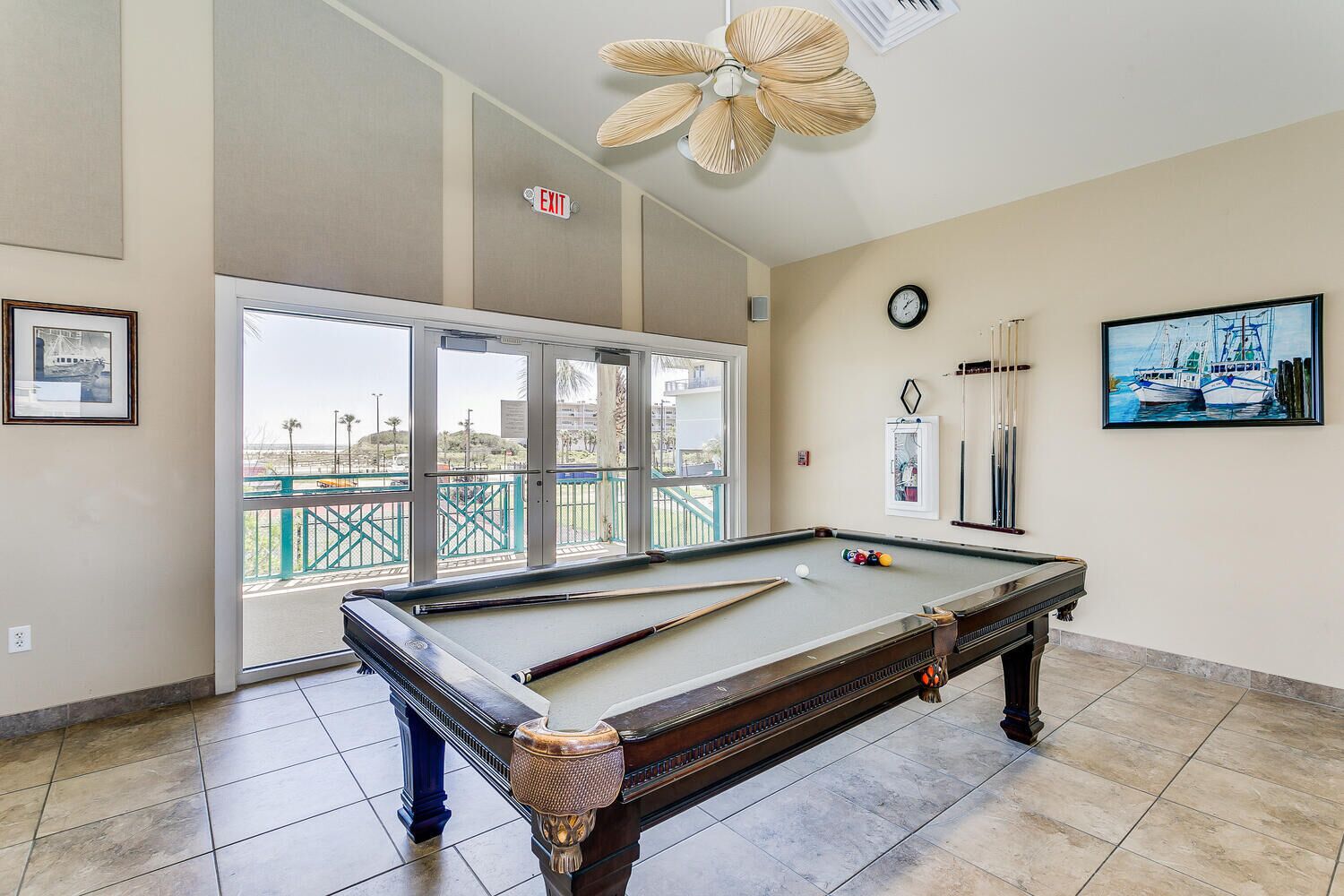 A pool table in a room with a large window, an exit sign, a clock, and a framed picture of boats.