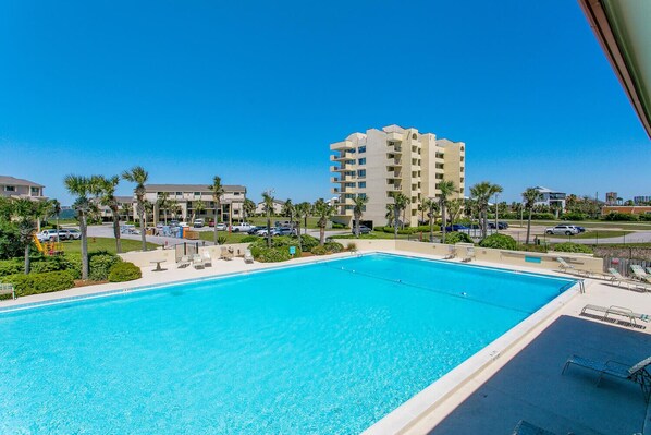 A clear blue pool with lounge chairs, palm trees, and a multi-story building in the background.