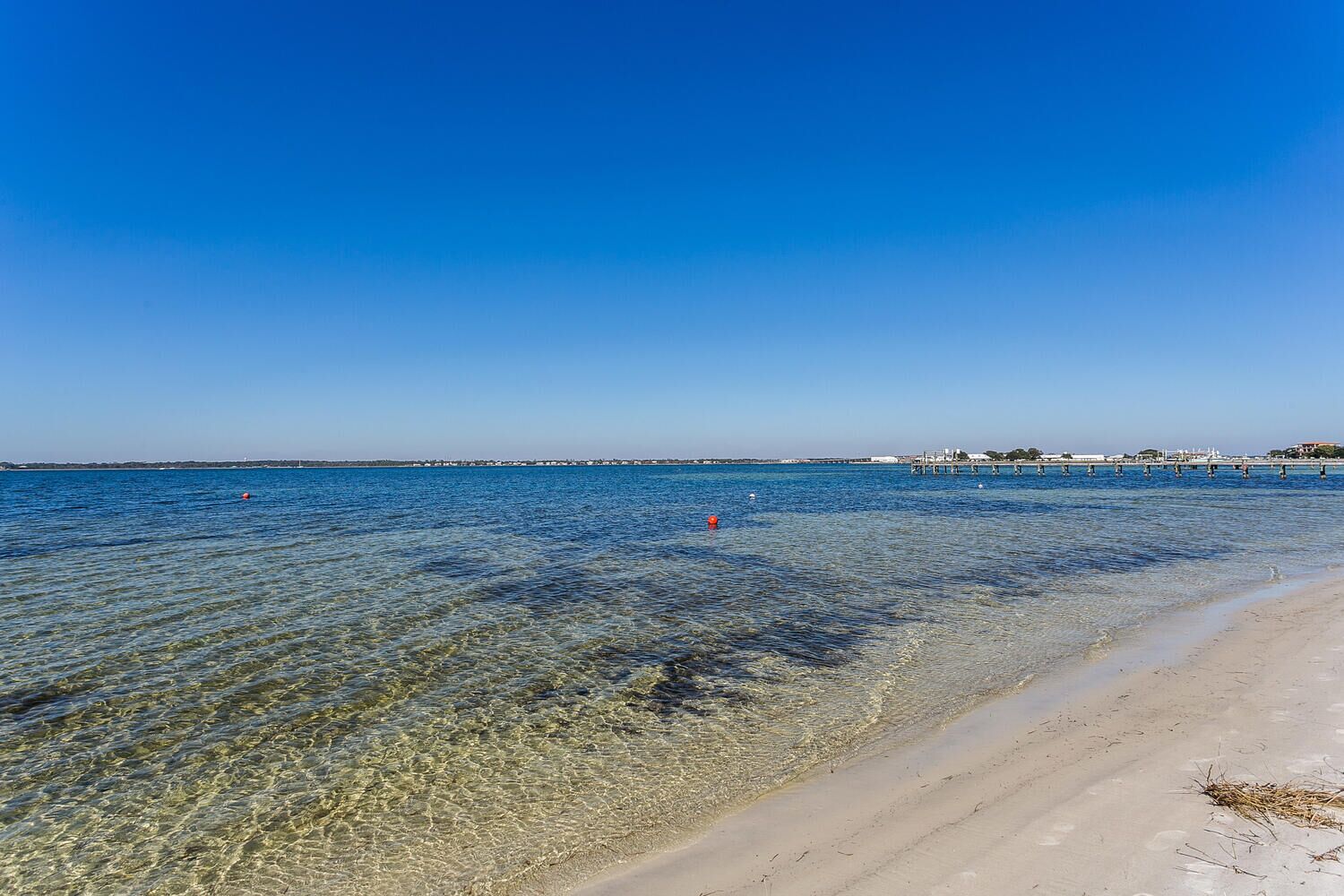 A beach with clear shallow waters, a sandy shore, and a distant pier with buildings.