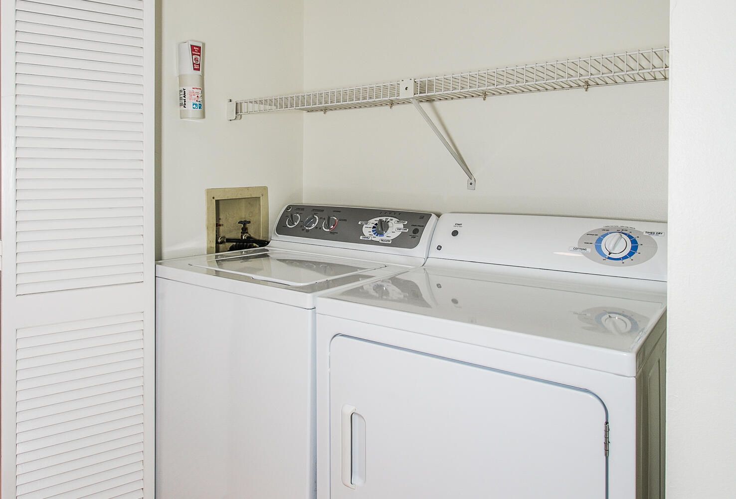 A laundry area with a front-loading washer and dryer, a metal shelf above the dryer, and a white wall.