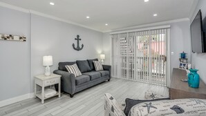Una sala de estar con un sofá gris, mesitas auxiliares blancas y un televisor montado en la pared.