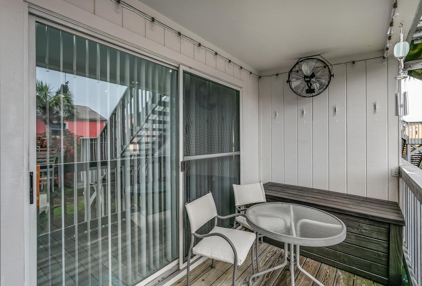 Ein Balkon mit Glastür, rundem Tisch, zwei Stühlen und einem Wandventilator.