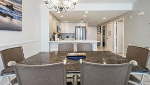 A dining area with a marble table, four upholstered chairs, a chandelier, and a kitchen in the background.