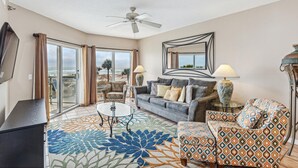 A spacious living room with a large window, a ceiling fan, a sofa, a patterned armchair, and a coffee table. - Great Location! Stunning Gulf Front Views at Emerald Isle! (Pensacola Beach)