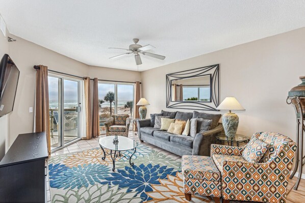 A spacious living room with a large window, a ceiling fan, a sofa, a patterned armchair, and a coffee table.