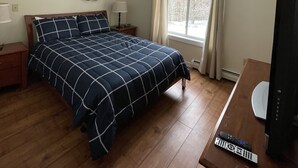A bedroom with a bed, wooden floor, a nightstand, a lamp, a window with curtains, and a TV stand with a remote control. - Cozy 2 BR Condo With Spectacular View of Mount Snow! (Dover)