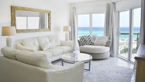 Amplia sala de estar con un gran ventanal que ofrece vistas al océano, un sofá blanco, una mesa de centro redonda y un sillón color beige.