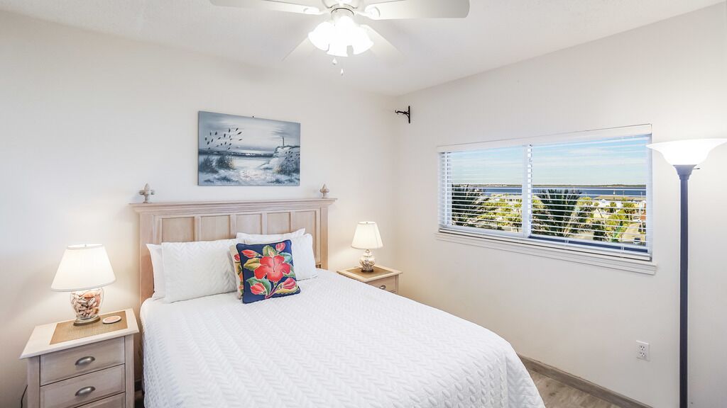 A bedroom with a bed, two bedside tables with lamps, a ceiling fan, a window with blinds, and a painting on the wall.